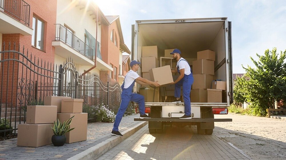 Professional movers loading cardboard moving boxes into a relocation truck outside a residential home during house shifting.