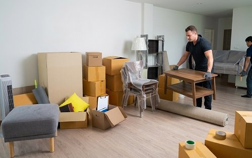 Mover carefully lifting a wooden coffee table in a room filled with protective-wrapped furniture and moving boxes