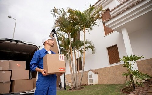 Uniformed mover carrying a fragile labeled cardboard box out of a delivery van in front of a modern residential home.