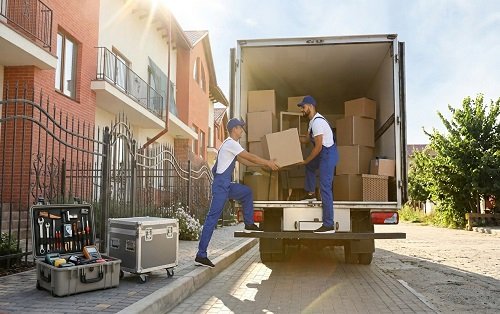 Two professional movers in blue uniforms loading cardboard boxes into an open moving truck outside a home