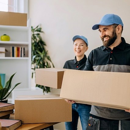 Two smiling movers in matching blue caps and uniforms carrying a cardboard box inside a modern home